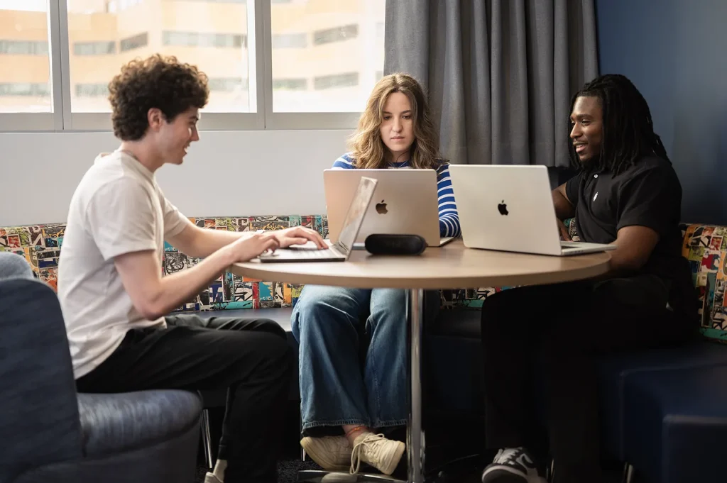 Three students working on laptops together at a round table in a lounge with colorful patterned seating. Photo by Matthew Nerber.