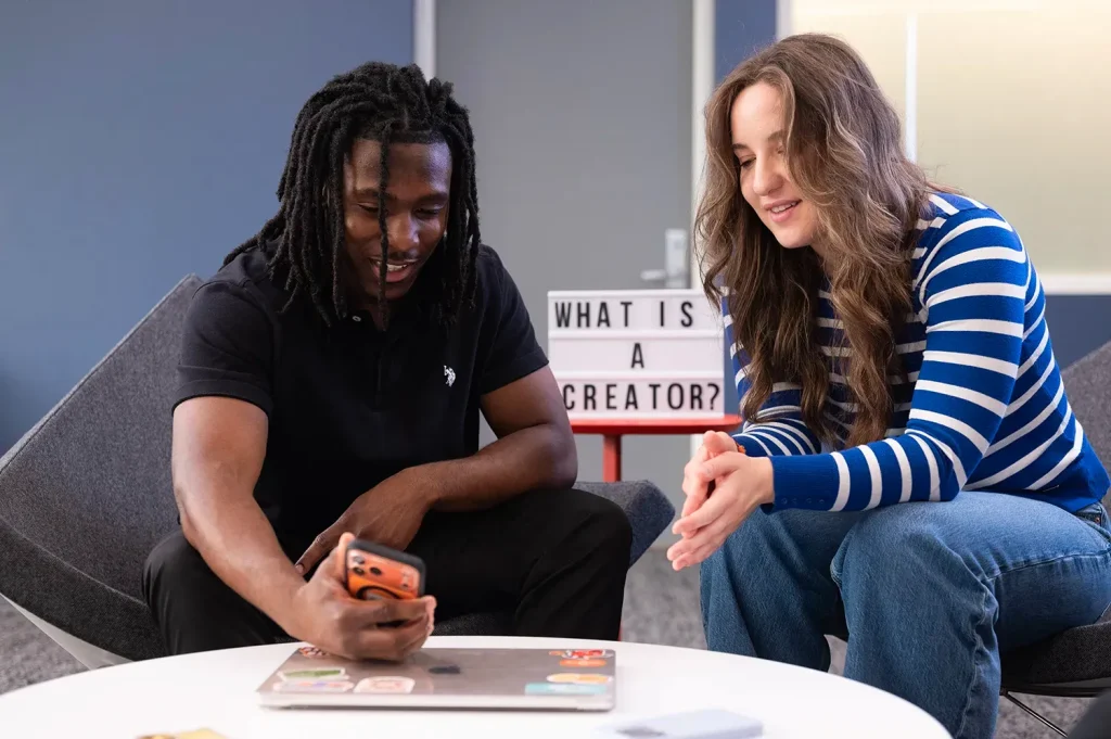 Two students looking at a smartphone together, seated near a sign reading 'What is a Creator?
